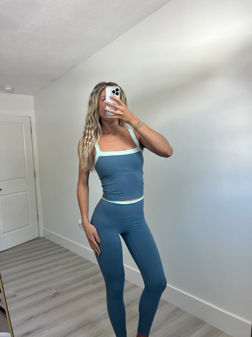 A woman takes a mirror selfie wearing a matching blue activewear set consisting of a scoop-neck tank top and high-waisted leggings with light blue trim, in a room with light walls and wood-look flooring.
