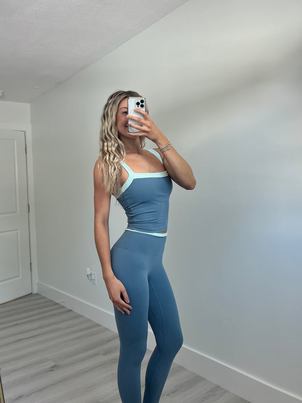 A woman takes a mirror selfie wearing a matching blue activewear set consisting of a scoop-neck tank top and high-waisted leggings with light blue trim, in a room with light walls and wood-look flooring.