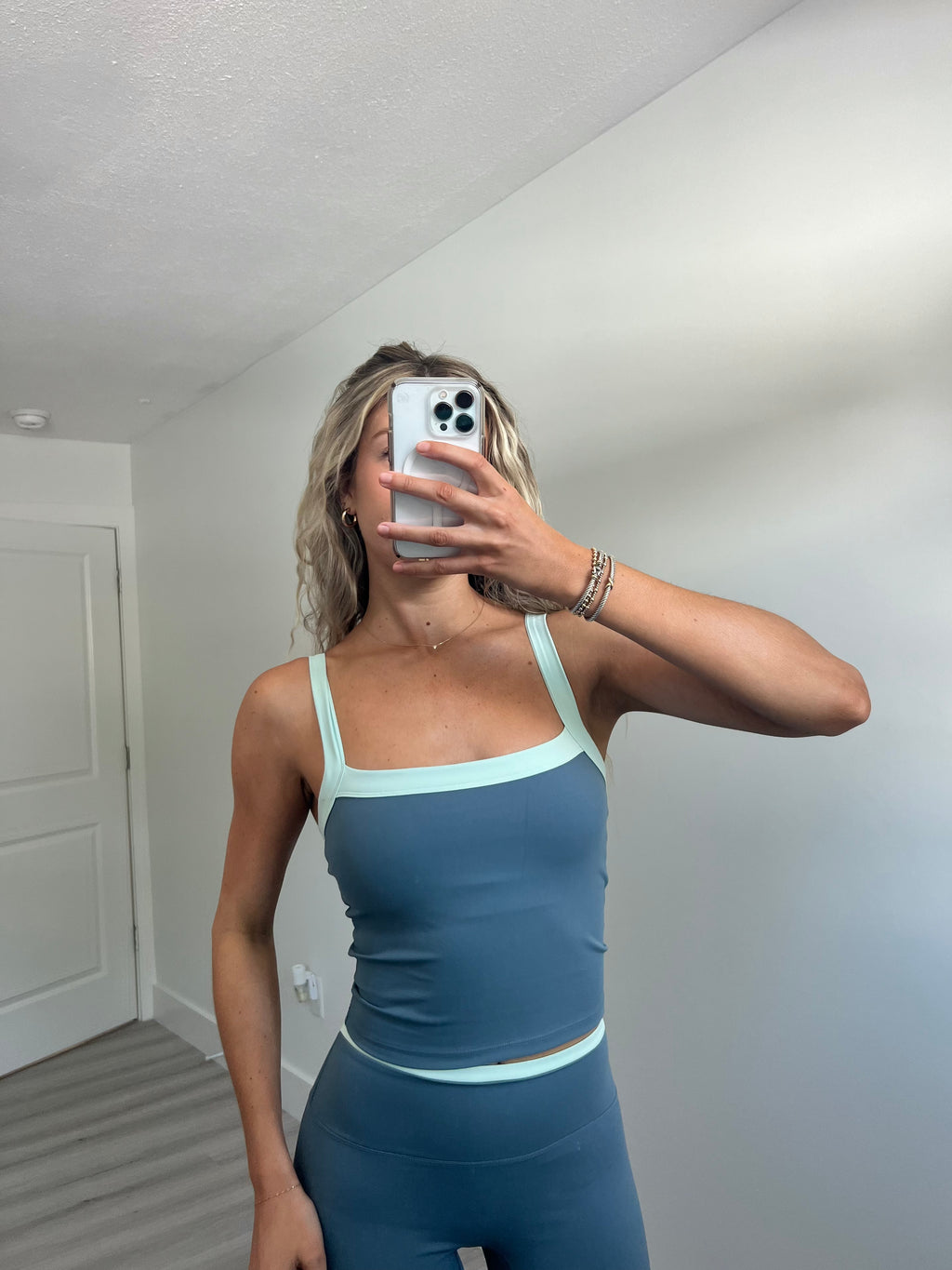 A woman takes a mirror selfie in a room wearing a matching blue athletic set consisting of a tank top bra and high-waisted leggings.