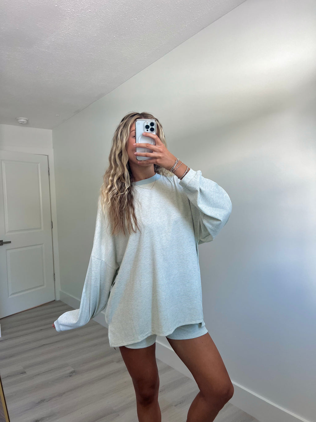 A woman takes a mirror selfie in a room wearing a light blue striped long-sleeve lounge set consisting of an oversized sweatshirt and matching shorts.