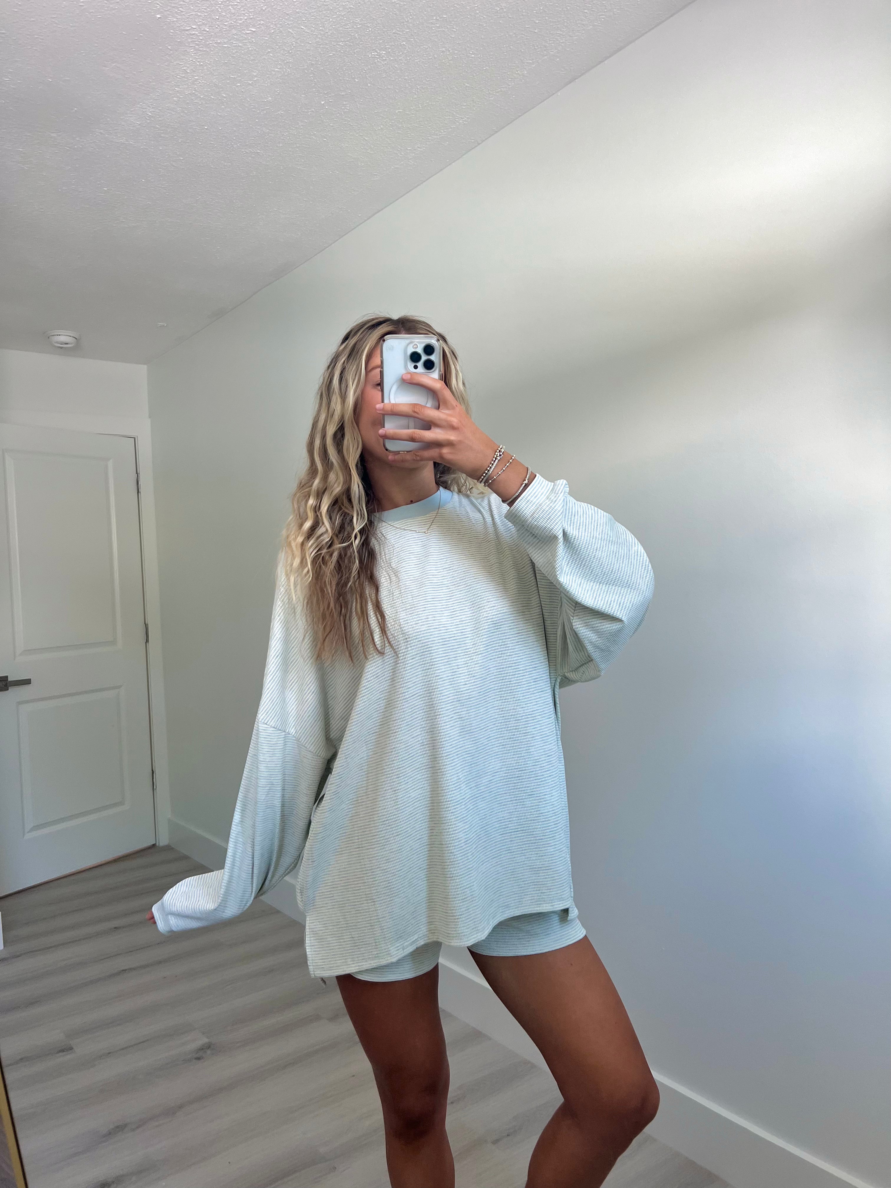 A woman takes a mirror selfie in a room wearing a light blue striped long-sleeve lounge set consisting of an oversized sweatshirt and matching shorts.