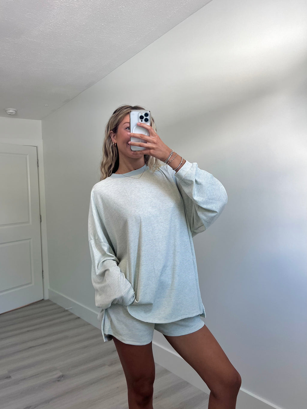 A woman takes a mirror selfie in a room wearing a light blue striped long-sleeve lounge set consisting of an oversized sweatshirt and matching shorts.
