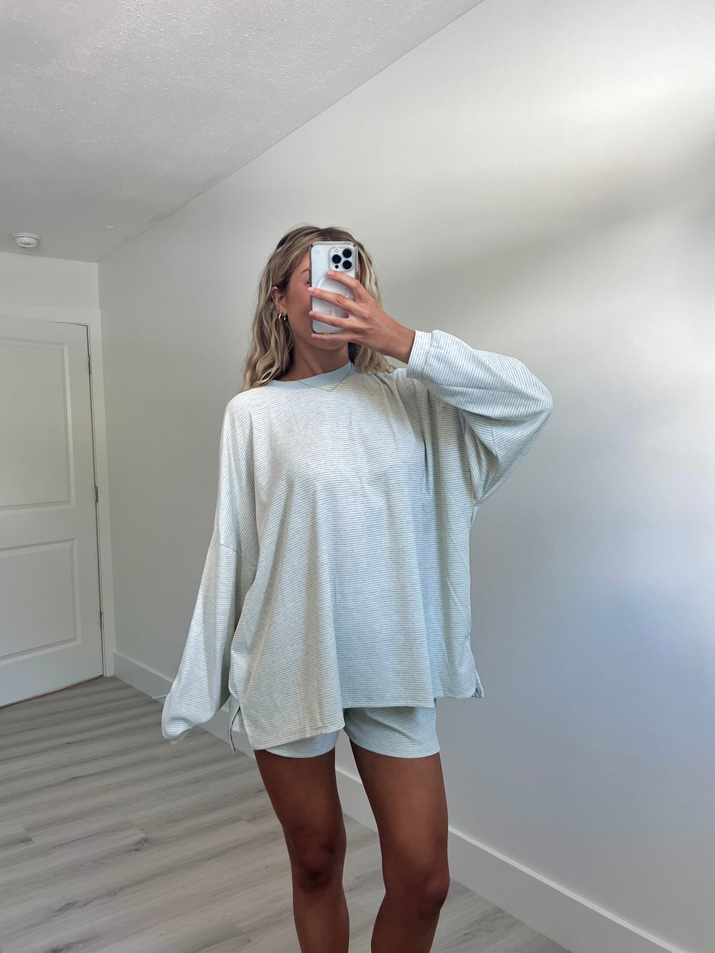 A woman takes a mirror selfie in a room wearing a light blue striped long-sleeve lounge set consisting of an oversized sweatshirt and matching shorts.