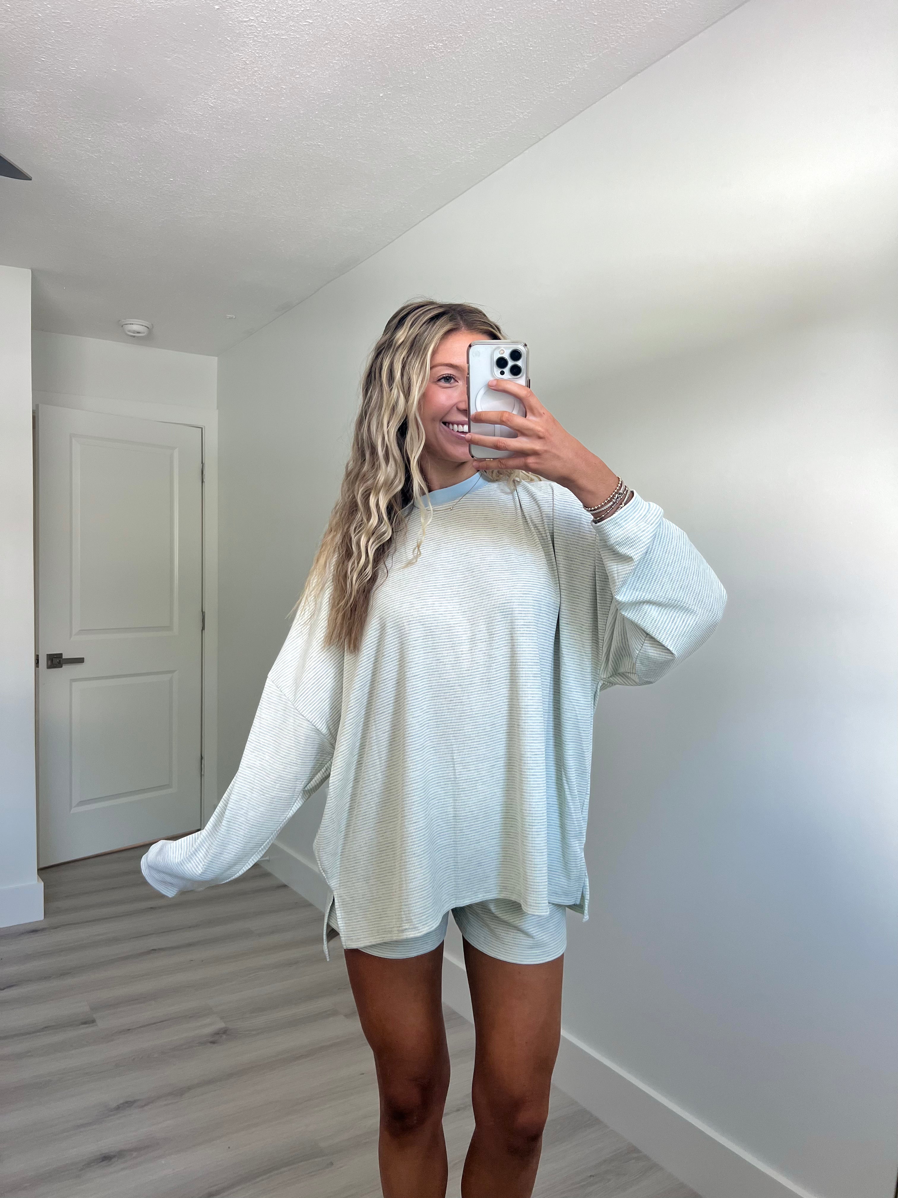 A woman takes a mirror selfie in a room wearing a light blue striped long-sleeve lounge set consisting of an oversized sweatshirt and matching shorts.
