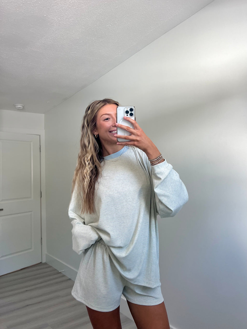 A woman takes a mirror selfie in a room wearing a light blue striped long-sleeve lounge set consisting of an oversized sweatshirt and matching shorts.