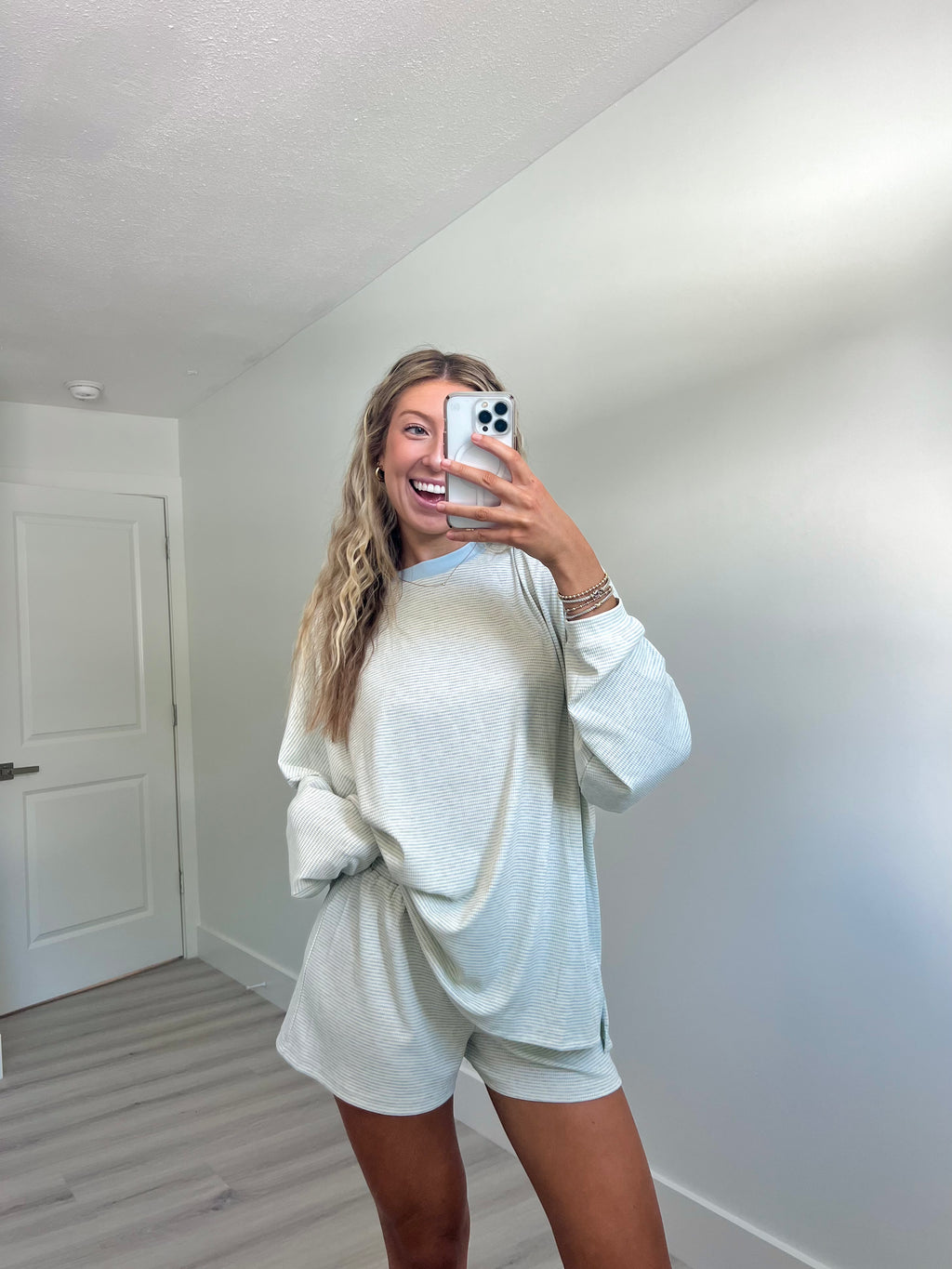 A woman takes a mirror selfie in a room wearing a light blue striped long-sleeve lounge set consisting of an oversized sweatshirt and matching shorts.