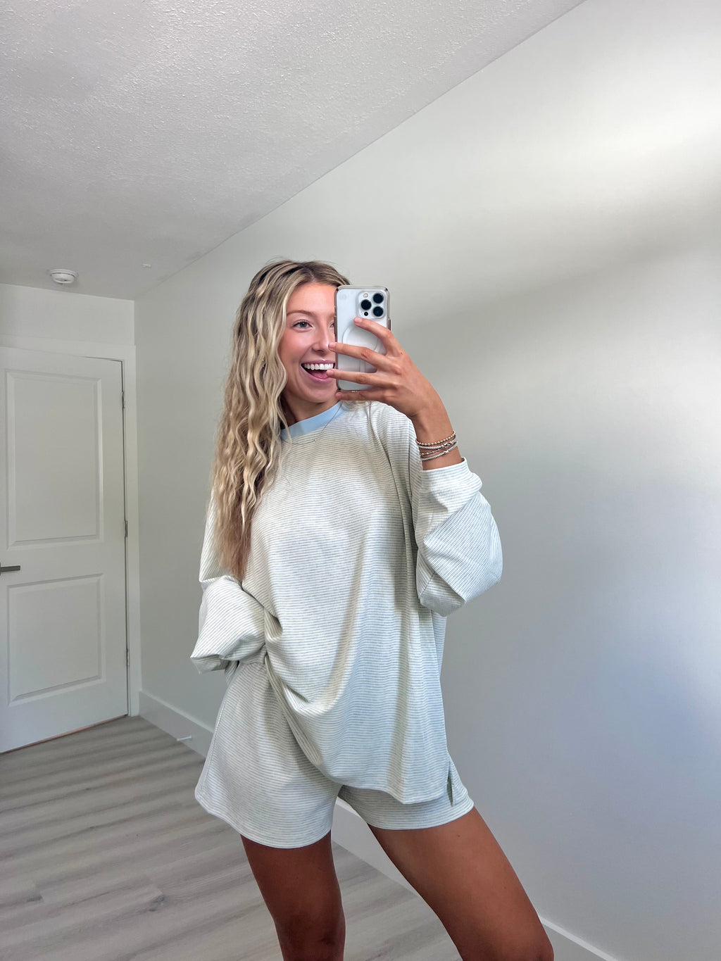 A woman takes a mirror selfie in a room wearing a light blue striped long-sleeve lounge set consisting of an oversized sweatshirt and matching shorts.