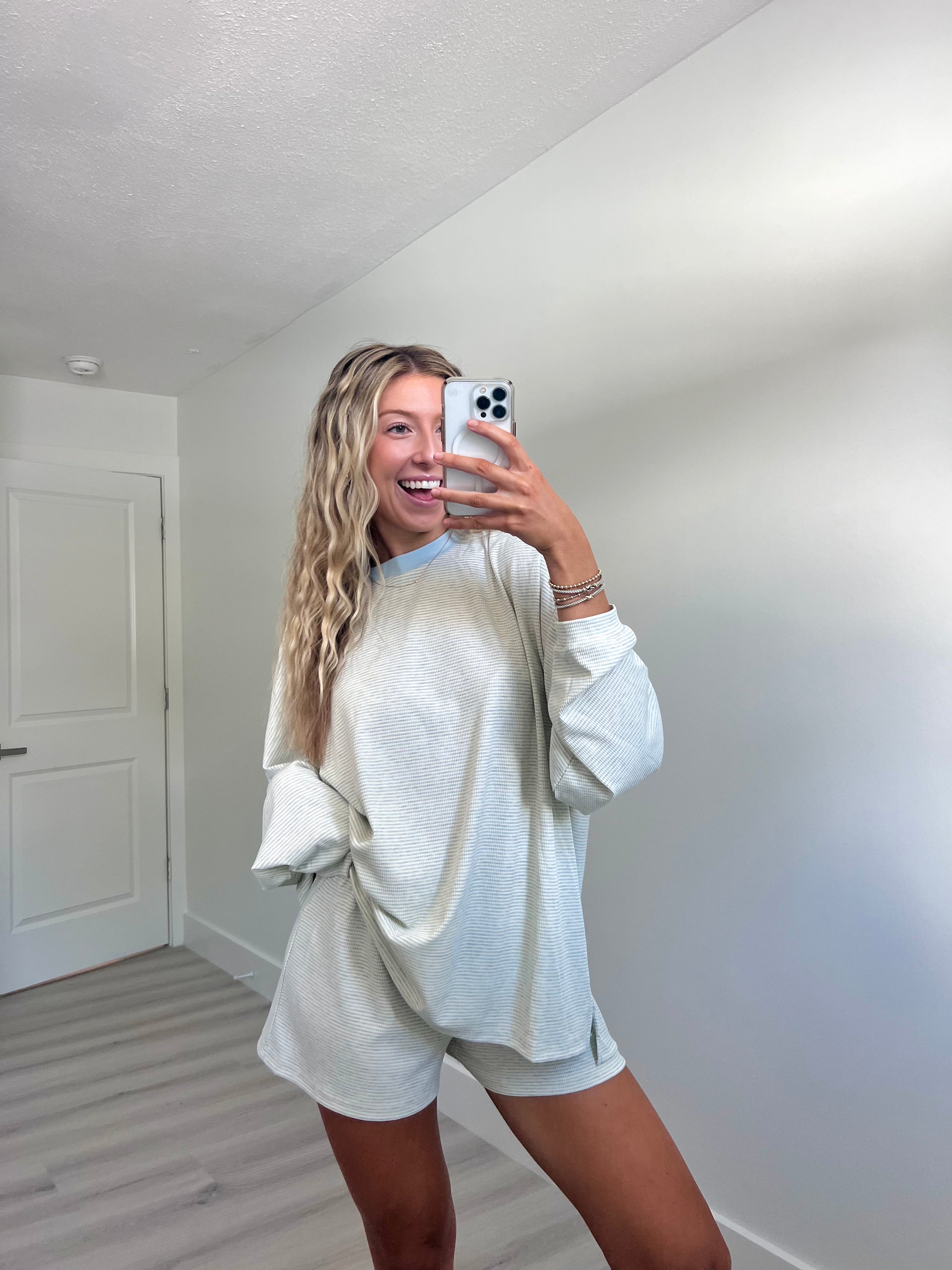 A woman takes a mirror selfie in a room wearing a light blue striped long-sleeve lounge set consisting of an oversized sweatshirt and matching shorts.