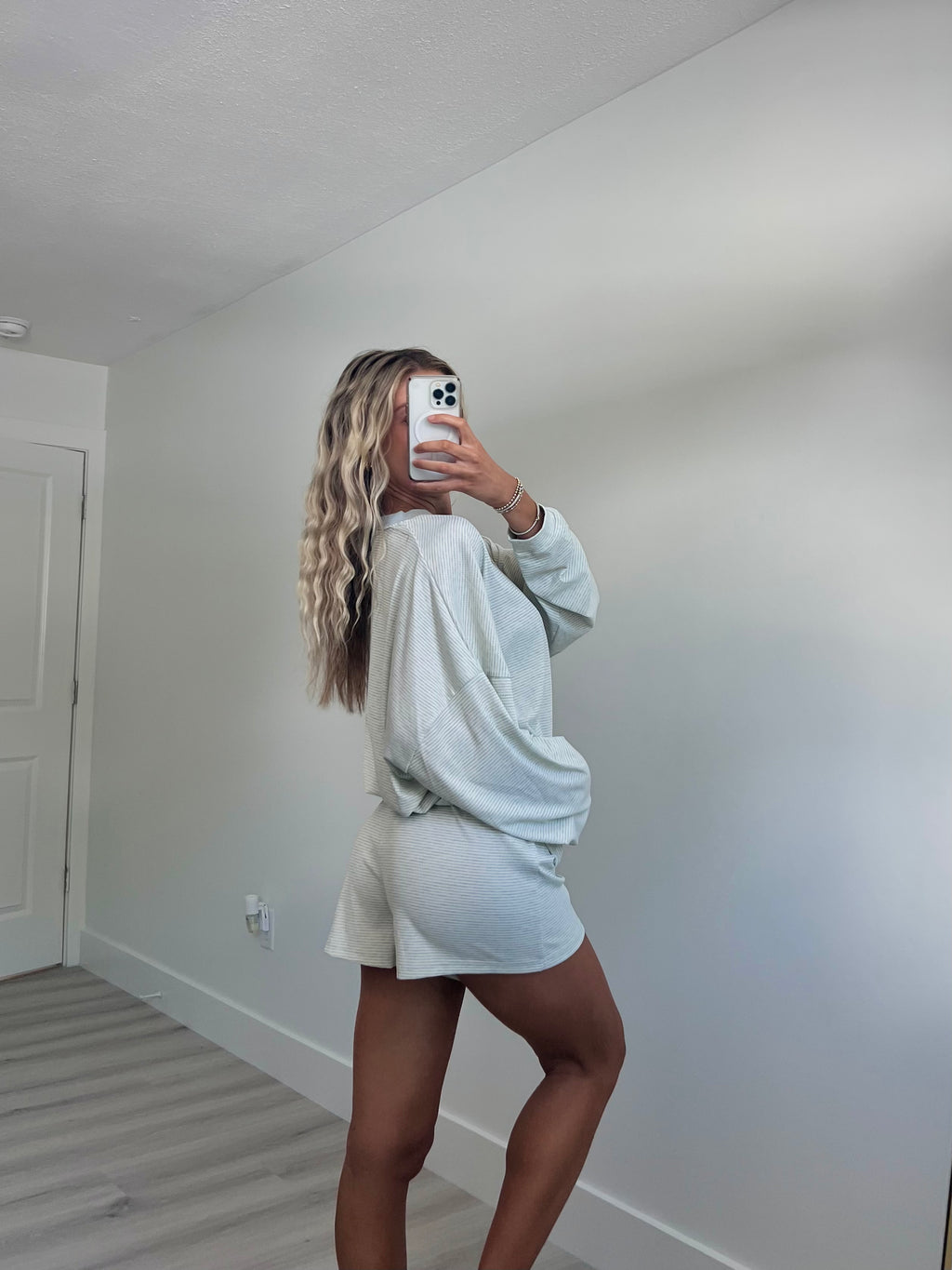 A woman takes a mirror selfie facing away from the camera in a room wearing a light blue striped long-sleeve lounge set consisting of an oversized sweatshirt and matching shorts.