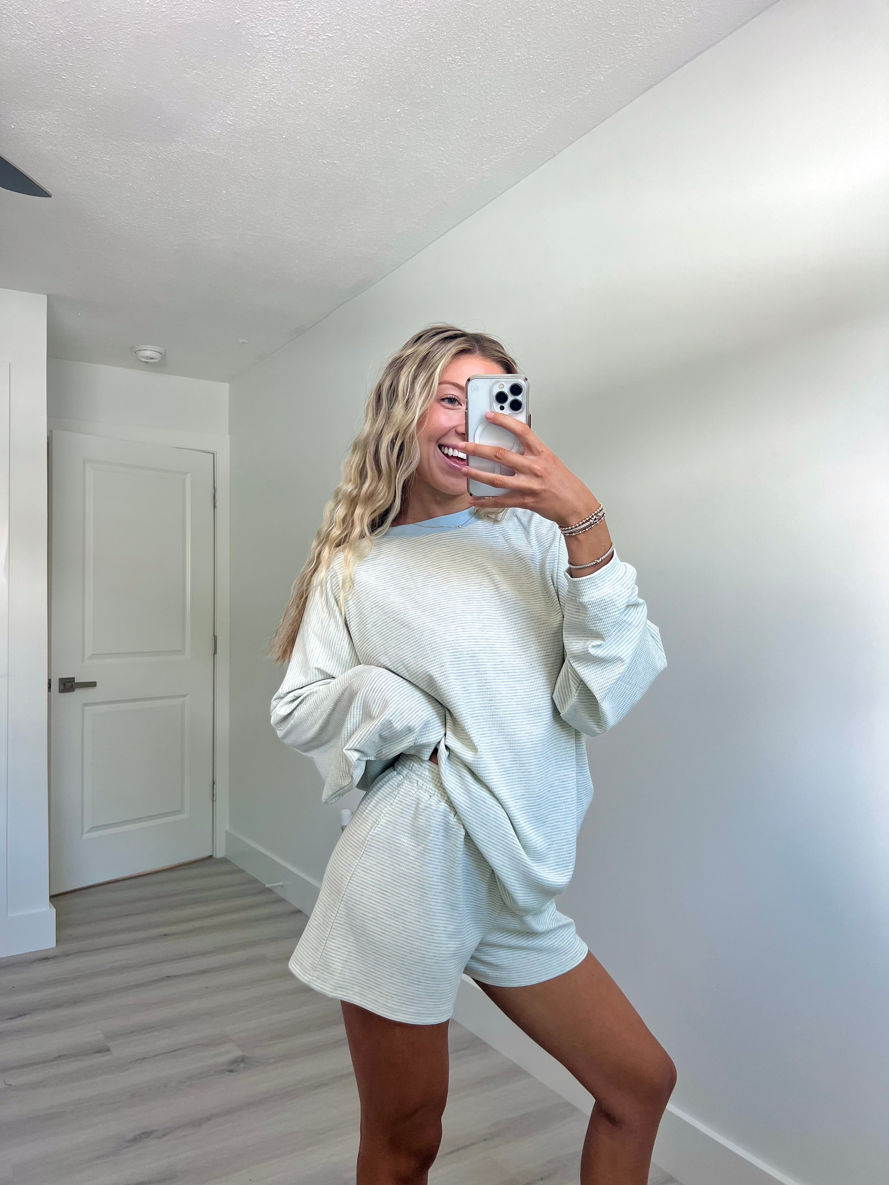 A woman takes a mirror selfie in a room wearing a light blue striped long-sleeve lounge set consisting of an oversized sweatshirt and matching shorts.