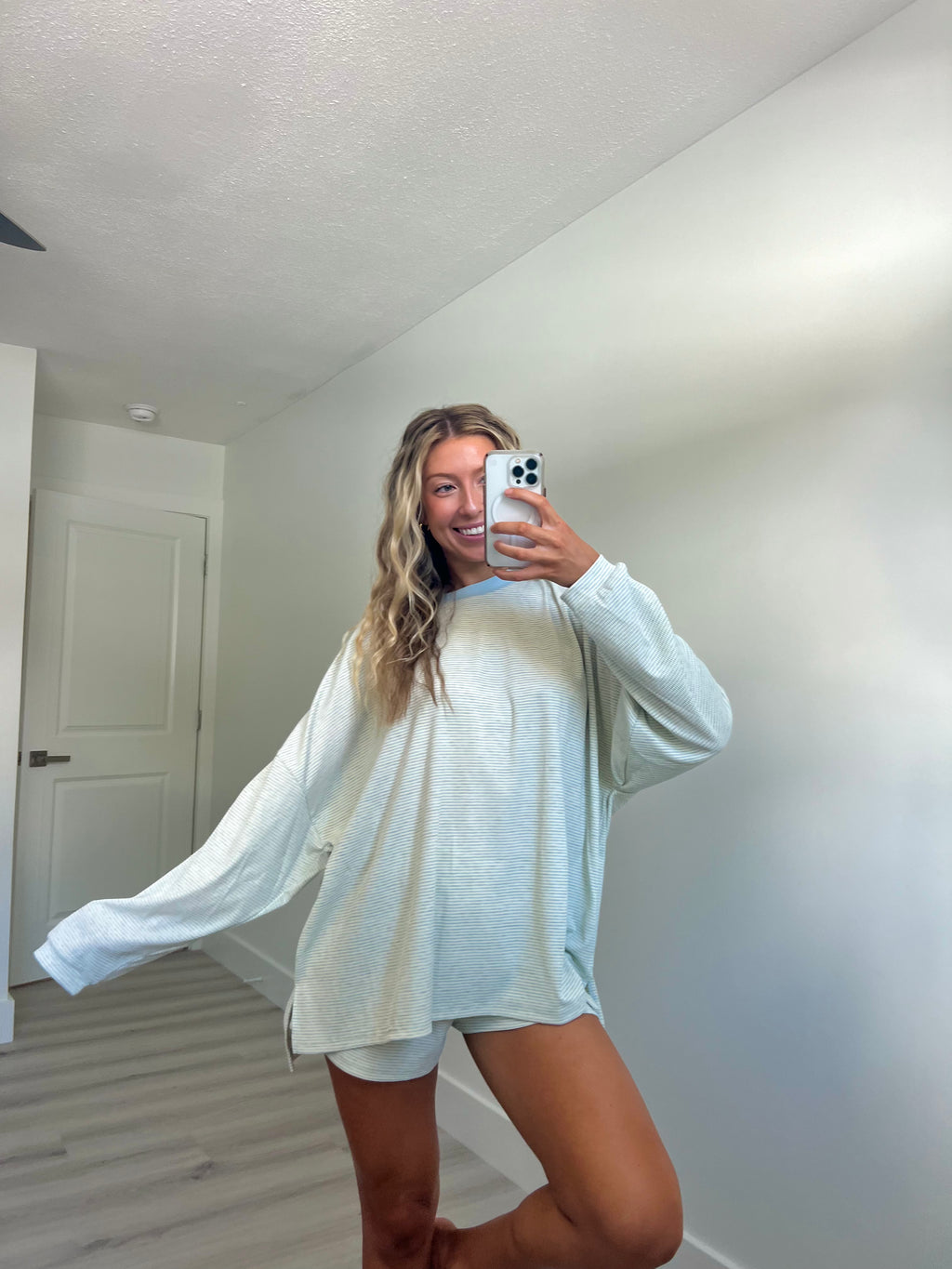 A woman takes a mirror selfie in a room wearing a light blue striped long-sleeve lounge set consisting of an oversized sweatshirt and matching shorts.