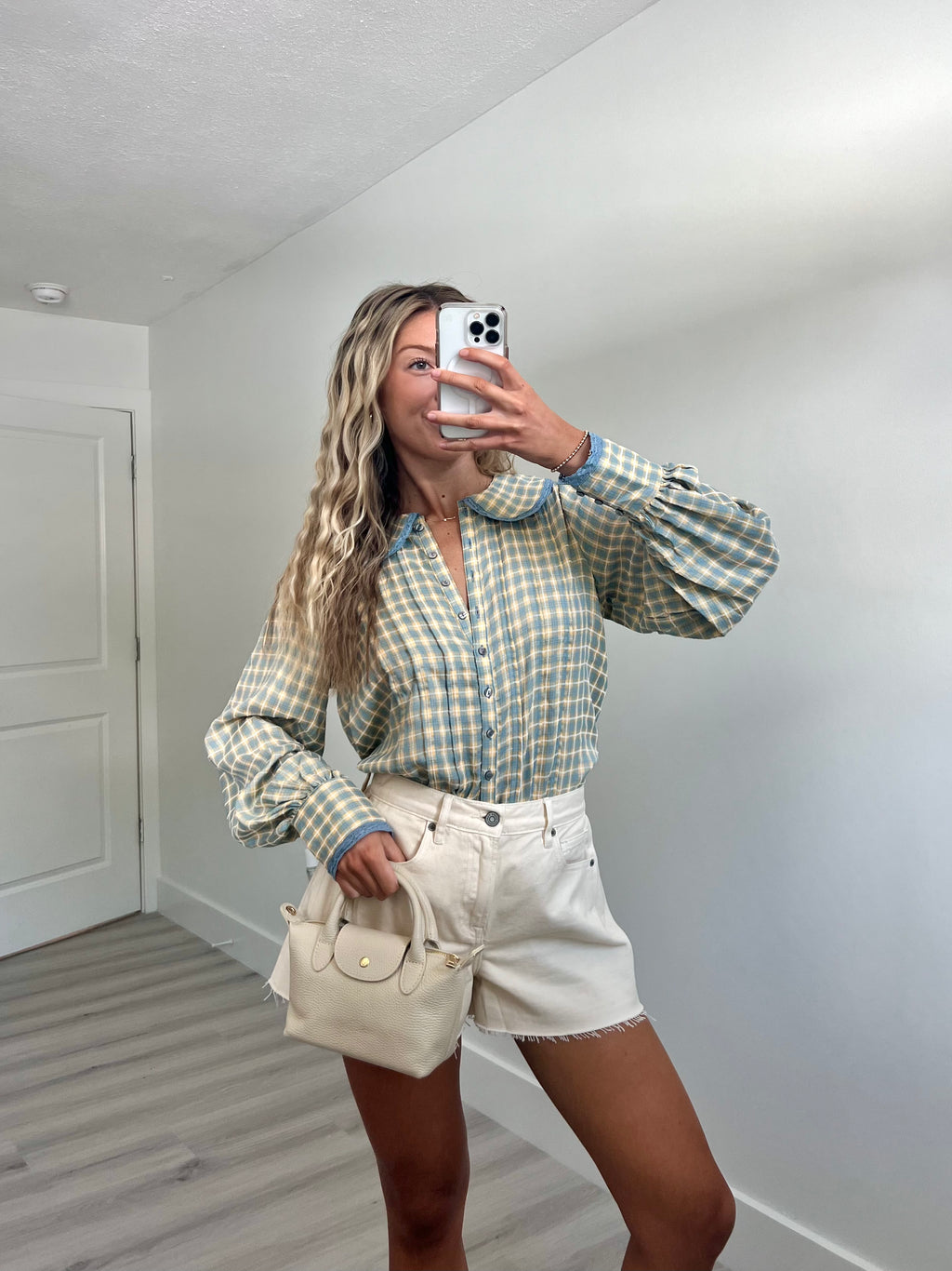 A woman takes a mirror selfie in a room, wearing a light blue and cream gingham long-sleeve button-up shirt, cream-colored denim shorts, and holding a beige handbag.