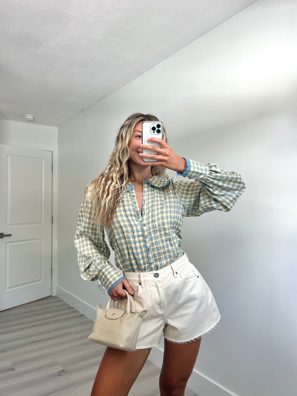 A woman takes a mirror selfie in a room, wearing a light blue and cream gingham long-sleeve button-up shirt, cream-colored denim shorts, and holding a beige handbag.