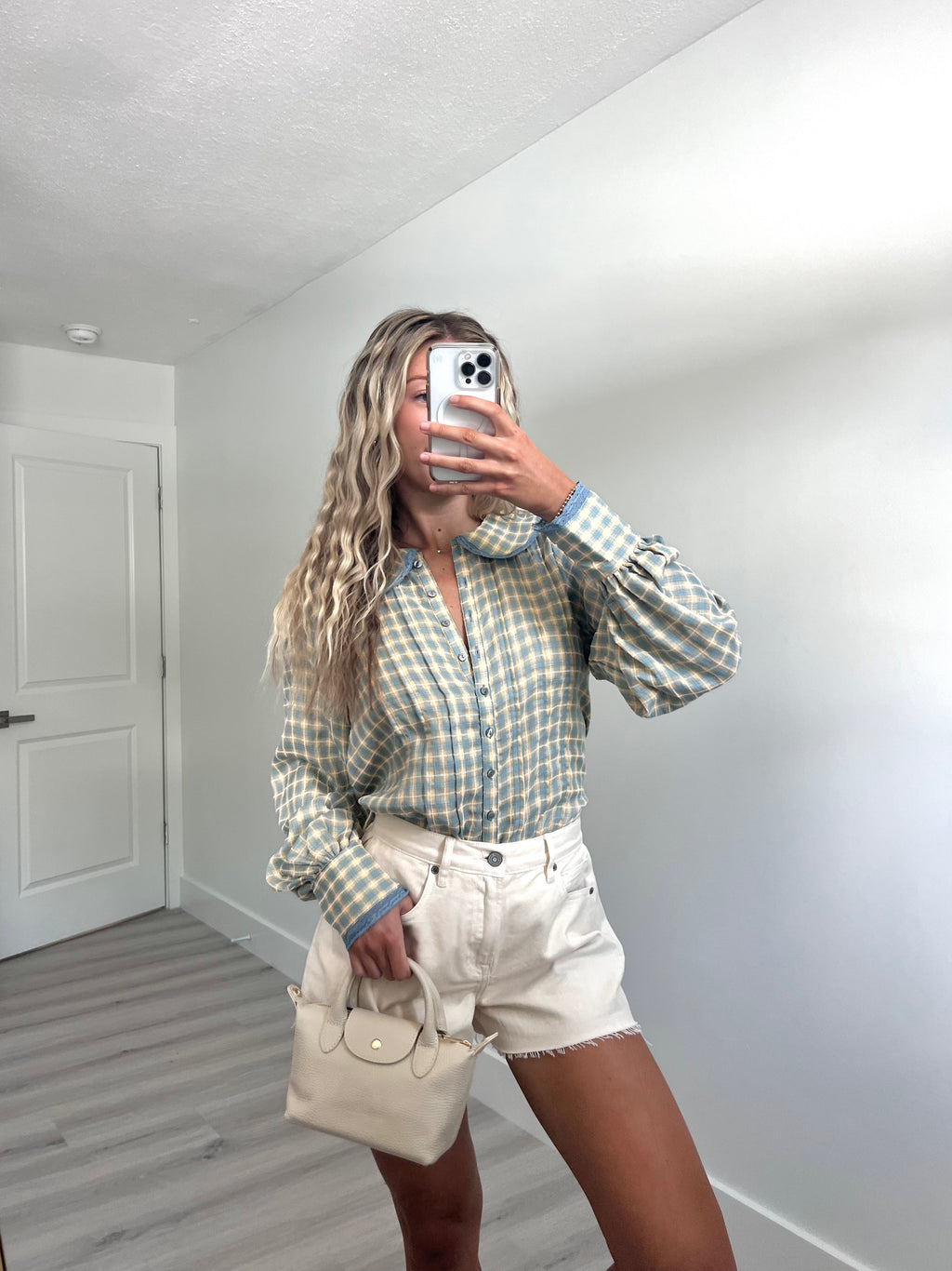 A woman takes a mirror selfie in a room, wearing a light blue and cream gingham long-sleeve button-up shirt, cream-colored denim shorts, and holding a beige handbag.