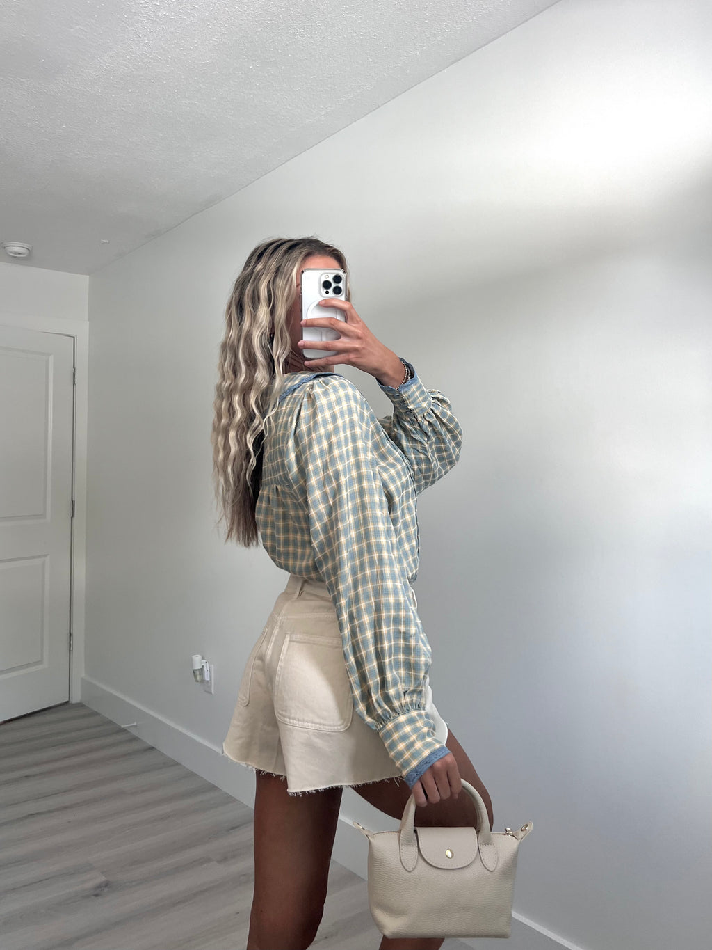 A woman takes a mirror selfie in a room, wearing a light blue and cream gingham long-sleeve button-up shirt, cream-colored denim shorts, and holding a beige handbag.