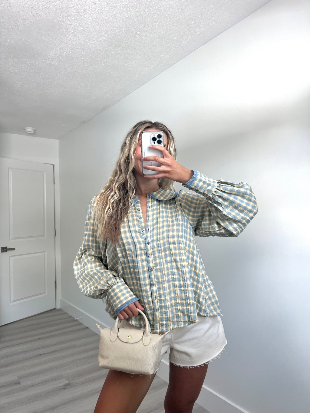 A woman takes a mirror selfie wearing a light blue and cream gingham long-sleeve button-up shirt with voluminous sleeves, paired with white shorts and holding a cream-colored handbag.
