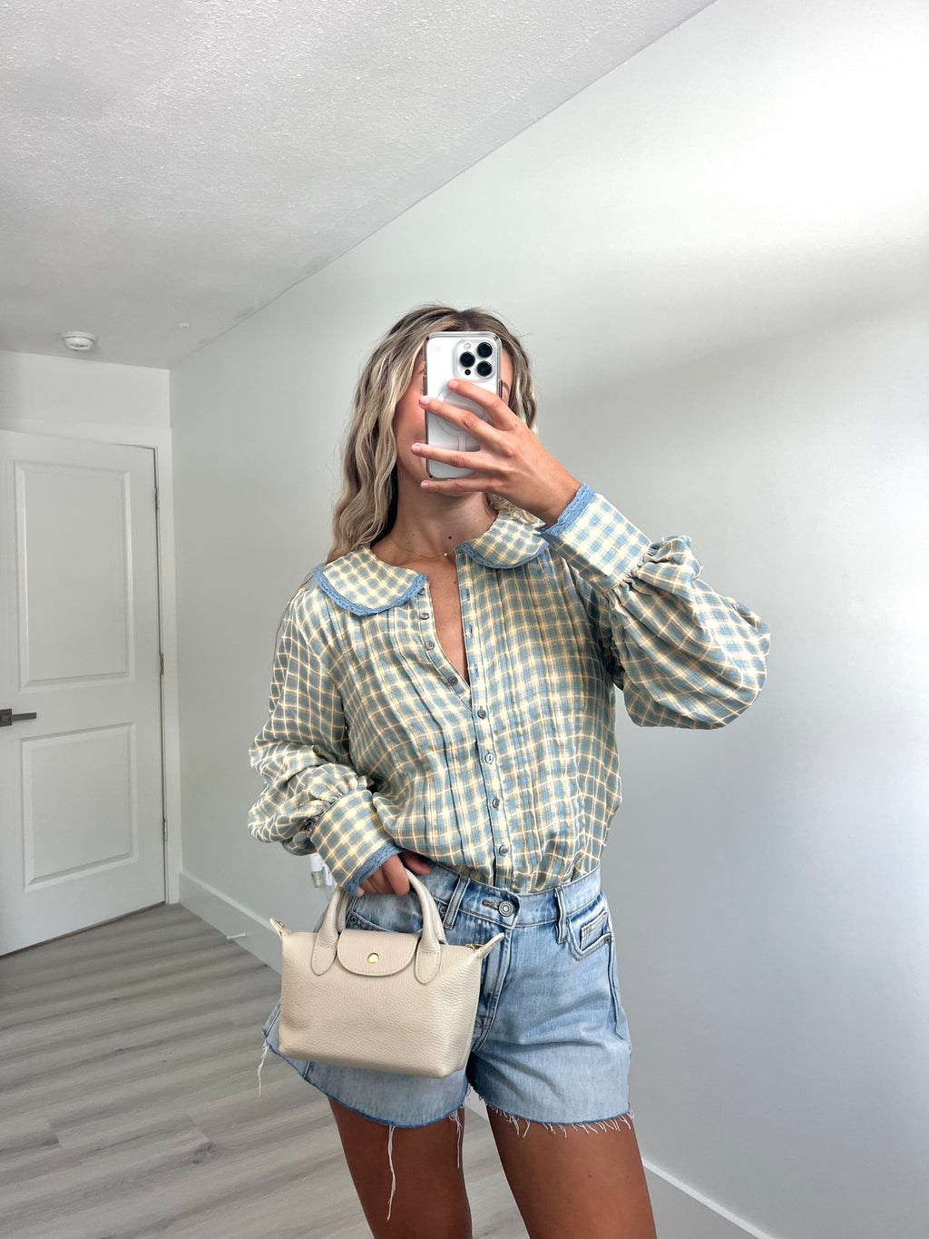 A woman takes a mirror selfie in a room, wearing a light blue and cream gingham long-sleeve button-up shirt, cream-colored denim shorts, and holding a beige handbag.