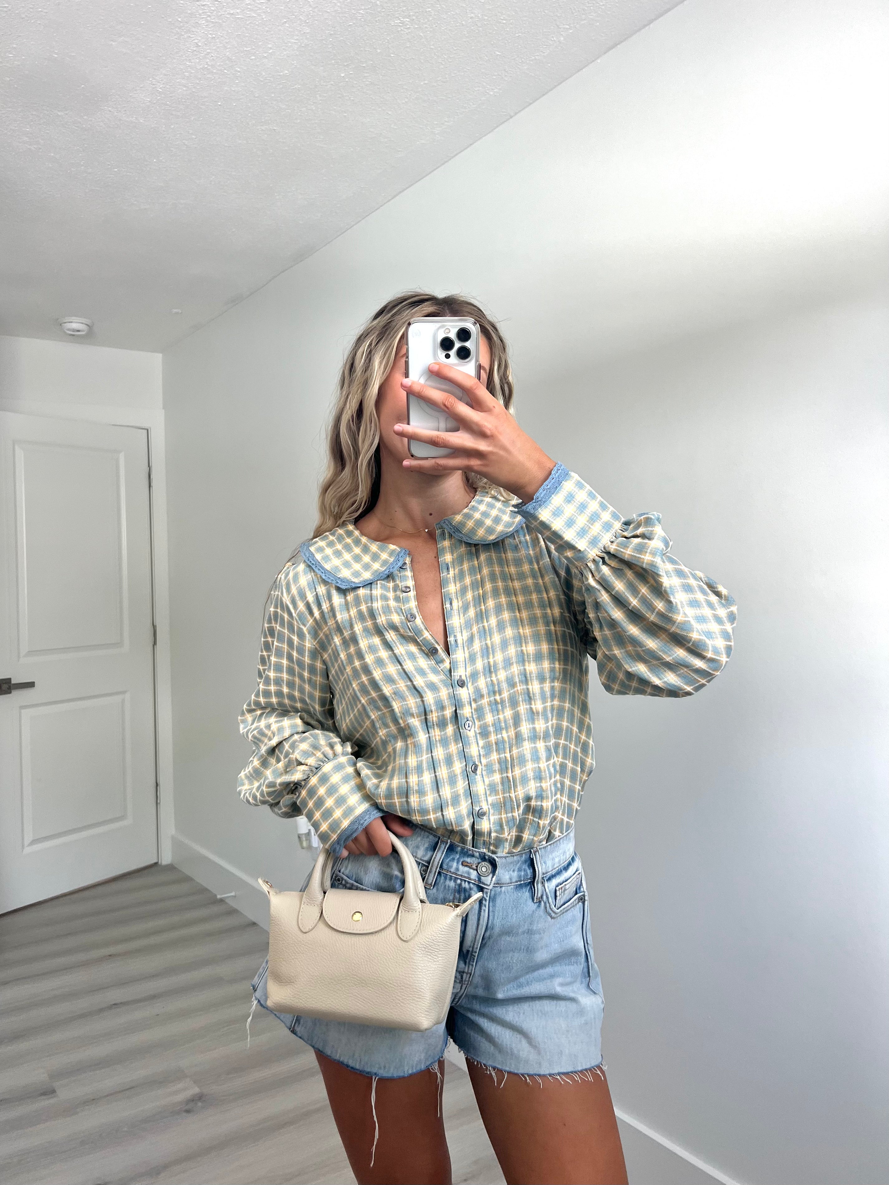 A woman takes a mirror selfie in a room, wearing a light blue and cream gingham long-sleeve button-up shirt, cream-colored denim shorts, and holding a beige handbag.