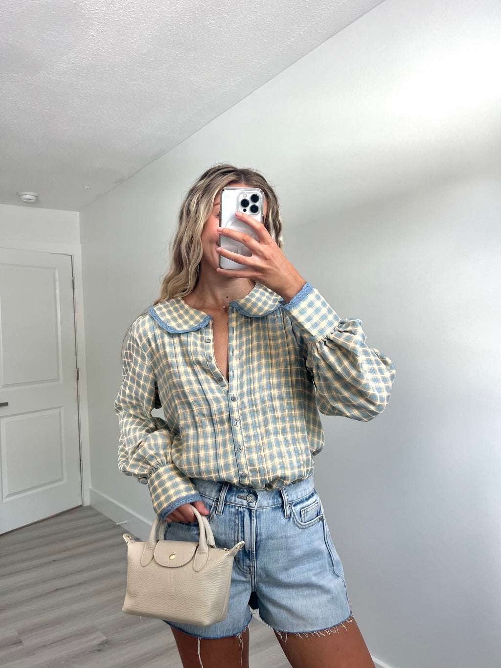 A woman takes a mirror selfie in a room, wearing a light blue and cream gingham long-sleeve button-up shirt, cream-colored denim shorts, and holding a beige handbag.