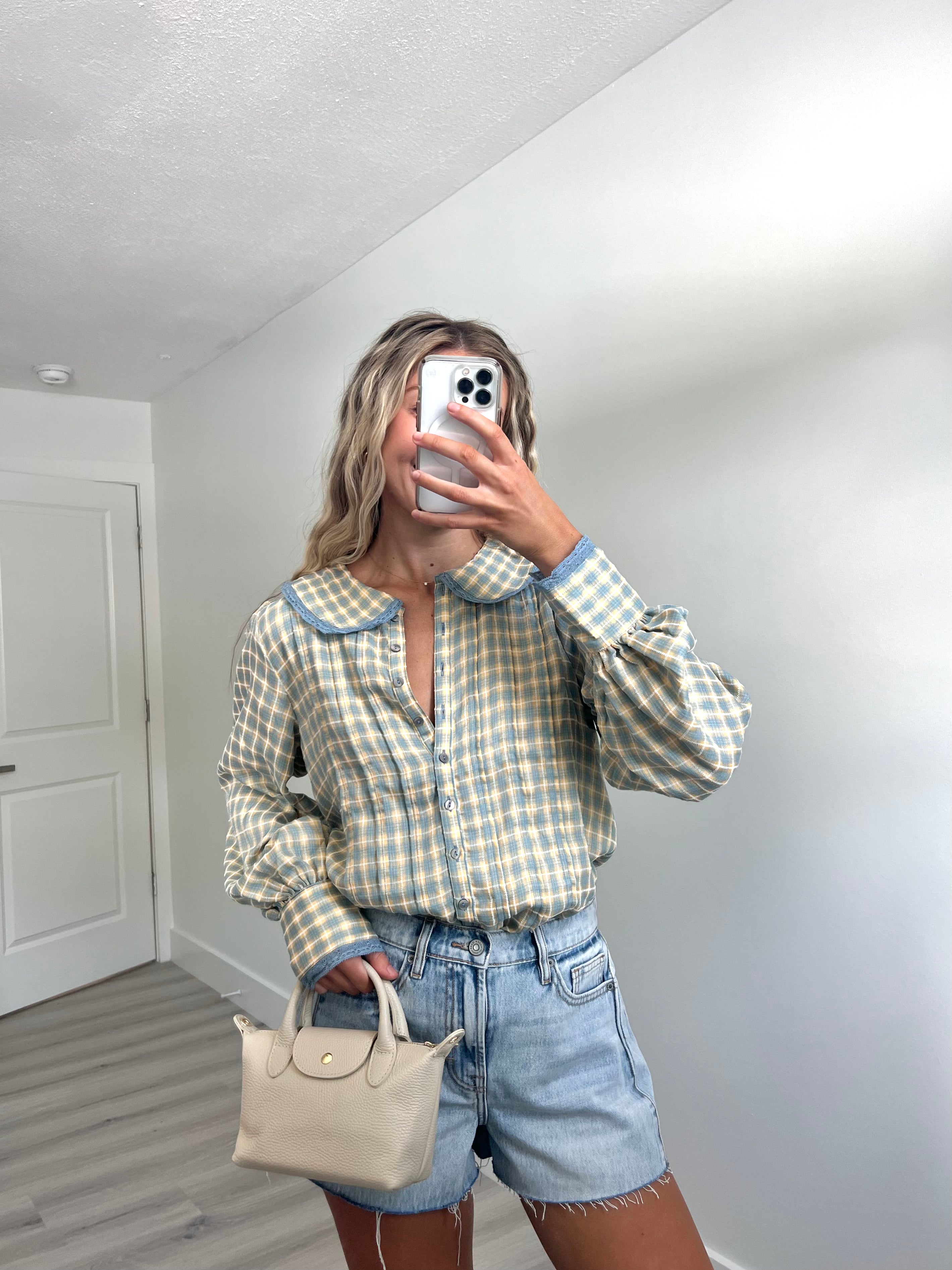 A woman takes a mirror selfie in a room, wearing a light blue and cream gingham long-sleeve button-up shirt, cream-colored denim shorts, and holding a beige handbag.