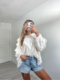 Woman taking a mirror selfie in a blank room with white walls and light wood floor, wearing a cream lace ruffle long sleeve top and denim shorts.