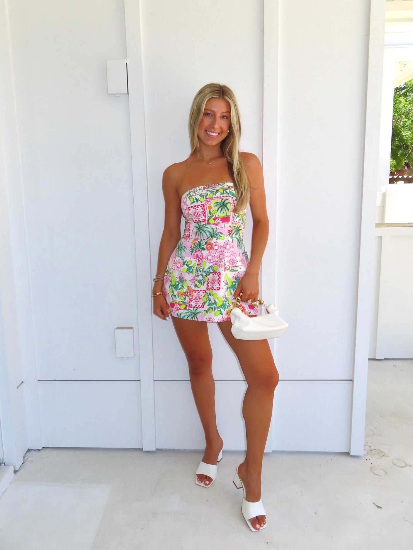 Woman wearing a floral strapless dress standing holding a white purse and standing on a white patio.