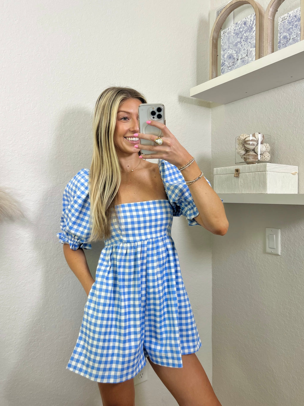 Woman wearing a babydoll blue and white gingham romper in a room with shelves and decor.