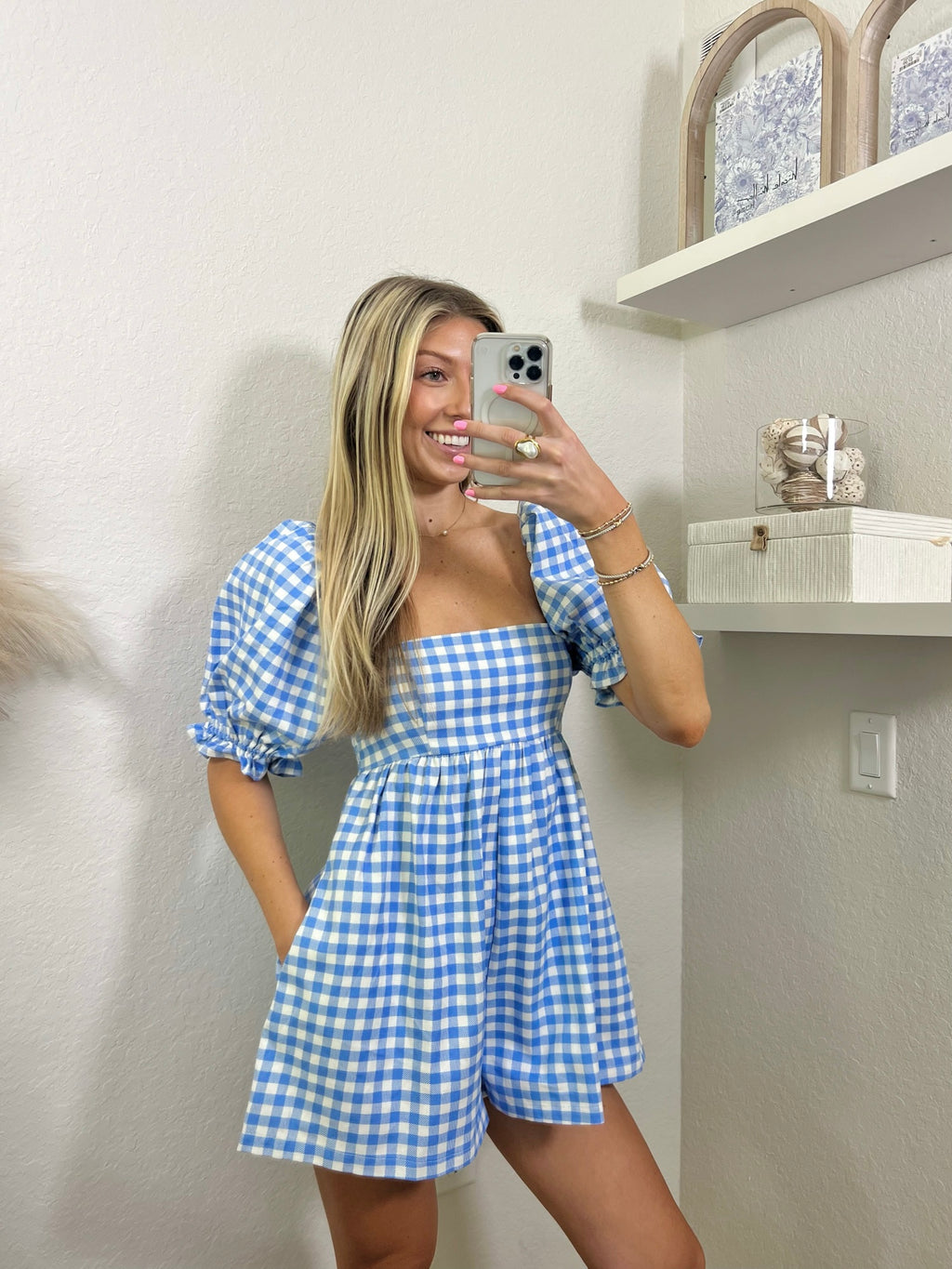 Woman wearing a babydoll blue and white gingham romper in a room with shelves and decor.