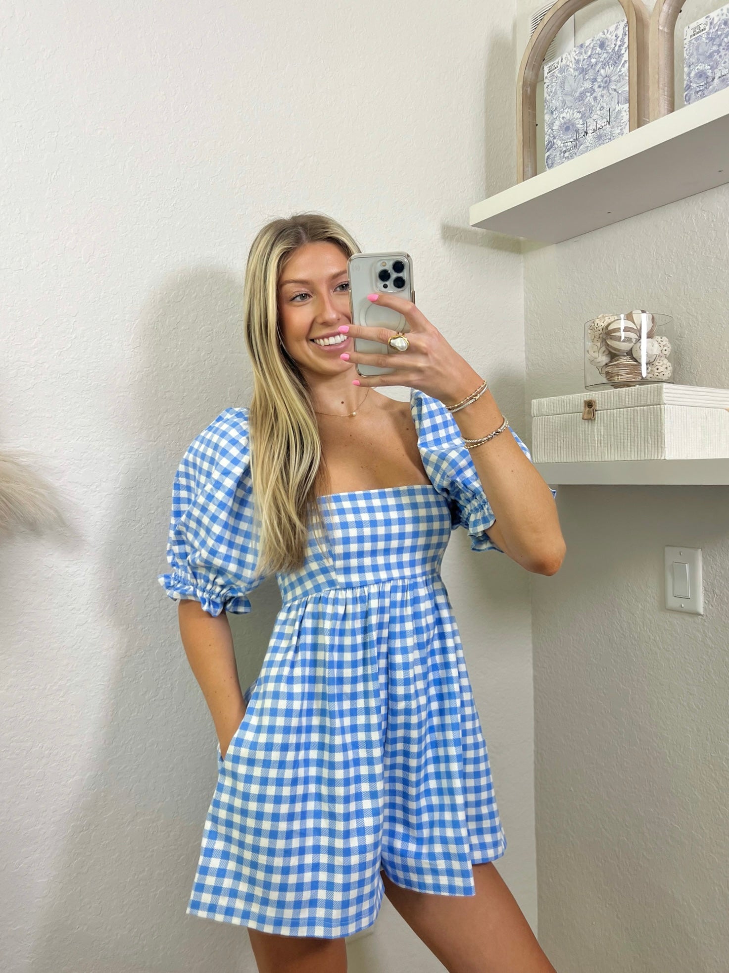 Woman wearing a babydoll blue and white gingham romper in a room with shelves and decor.