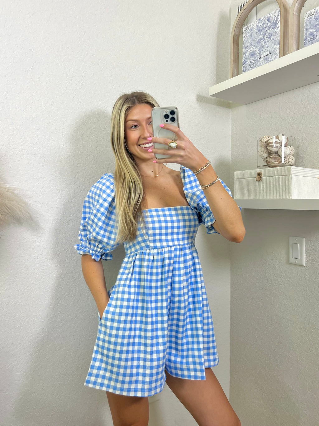 Woman wearing a babydoll blue and white gingham romper in a room with shelves and decor.
