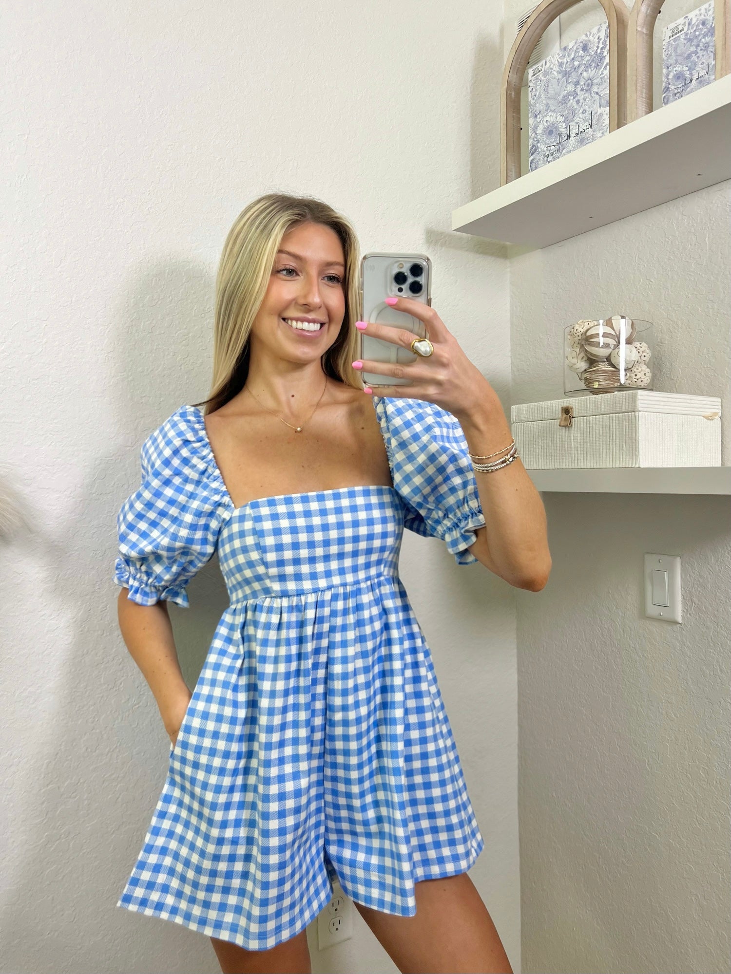 Woman wearing a babydoll blue and white gingham romper in a room with shelves and decor.