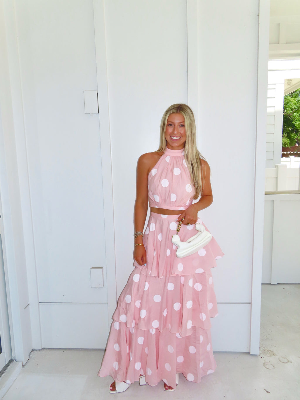 Woman wearing a pink polka dot skirt set holding a white clutch on a bright white patio.