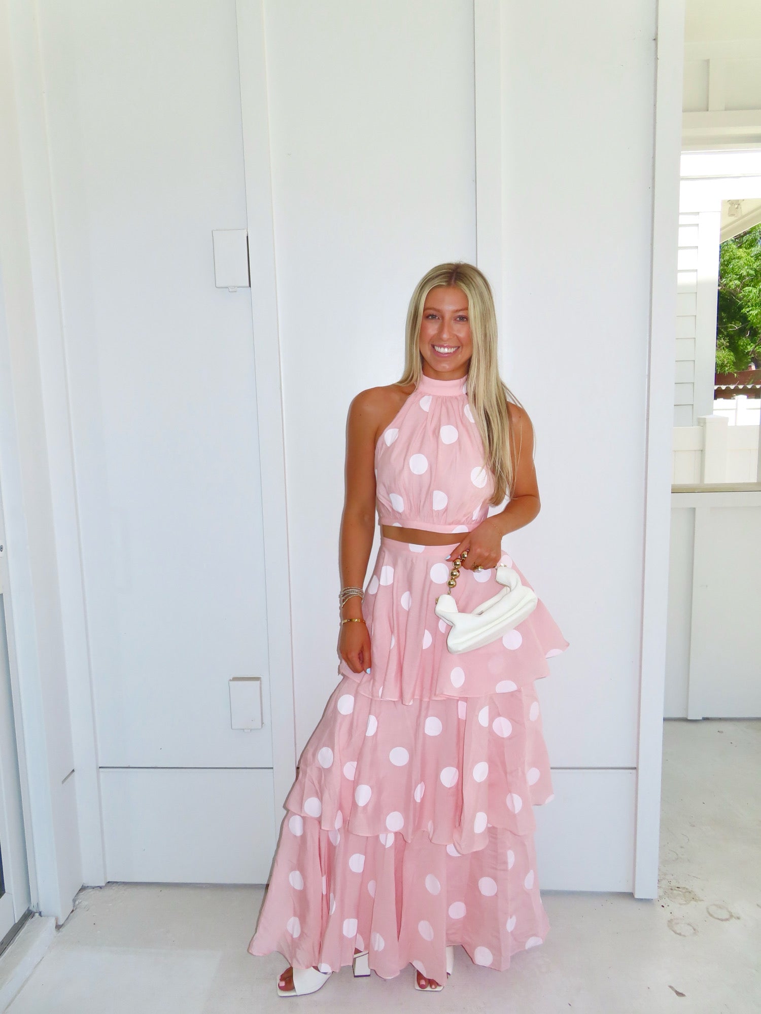 Woman wearing a pink polka dot skirt set holding a white clutch on a bright white patio.