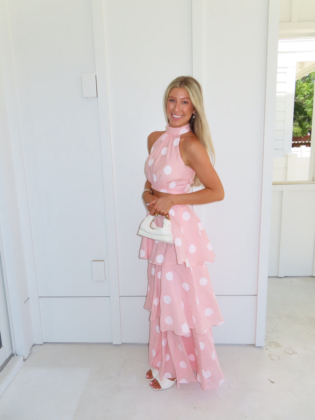 Woman wearing a pink polka dot skirt set holding a white clutch on a bright white patio.