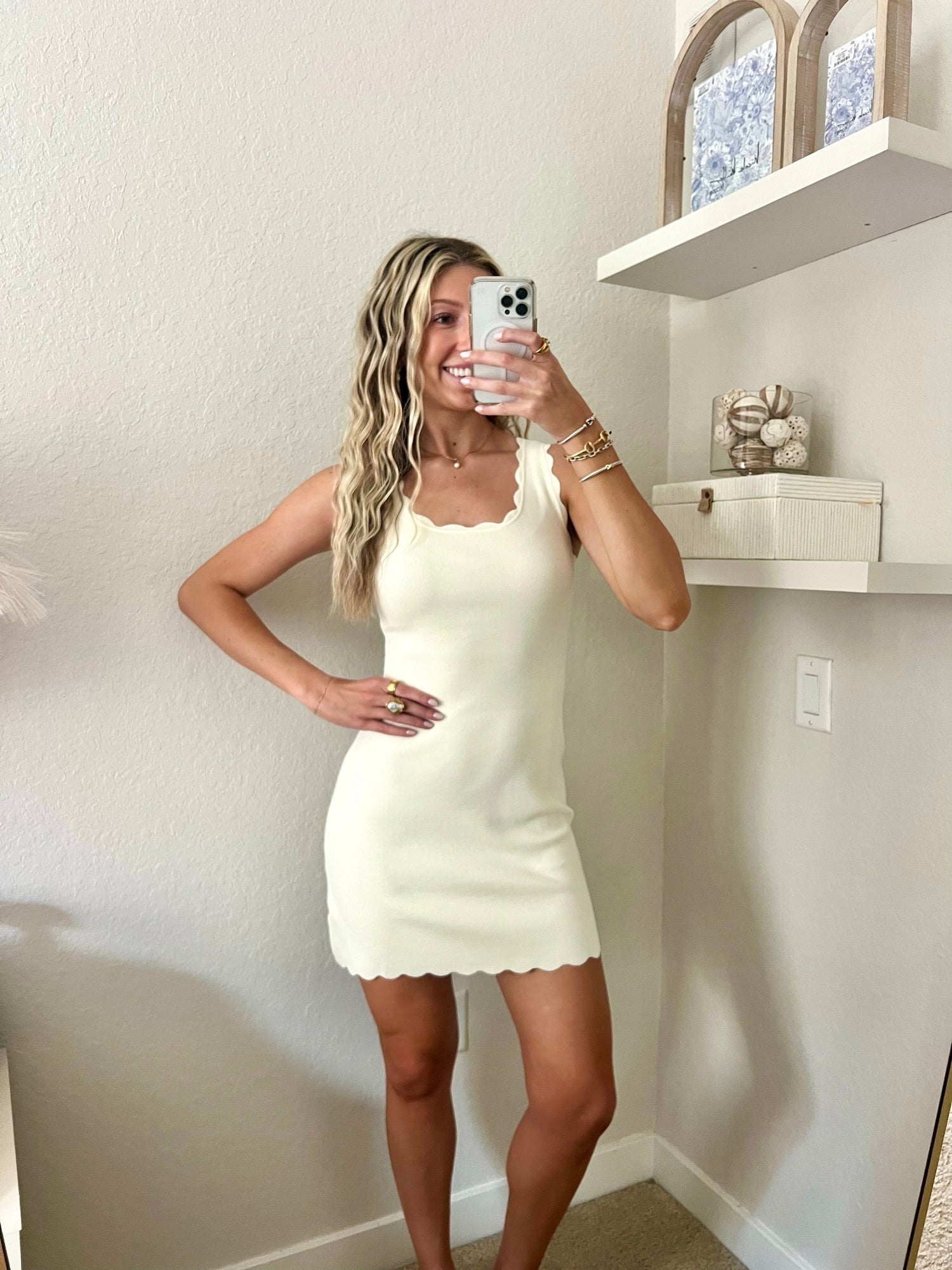 Woman taking a mirror selfie wearing an ivory dress in a room with a shelf and decorative items.