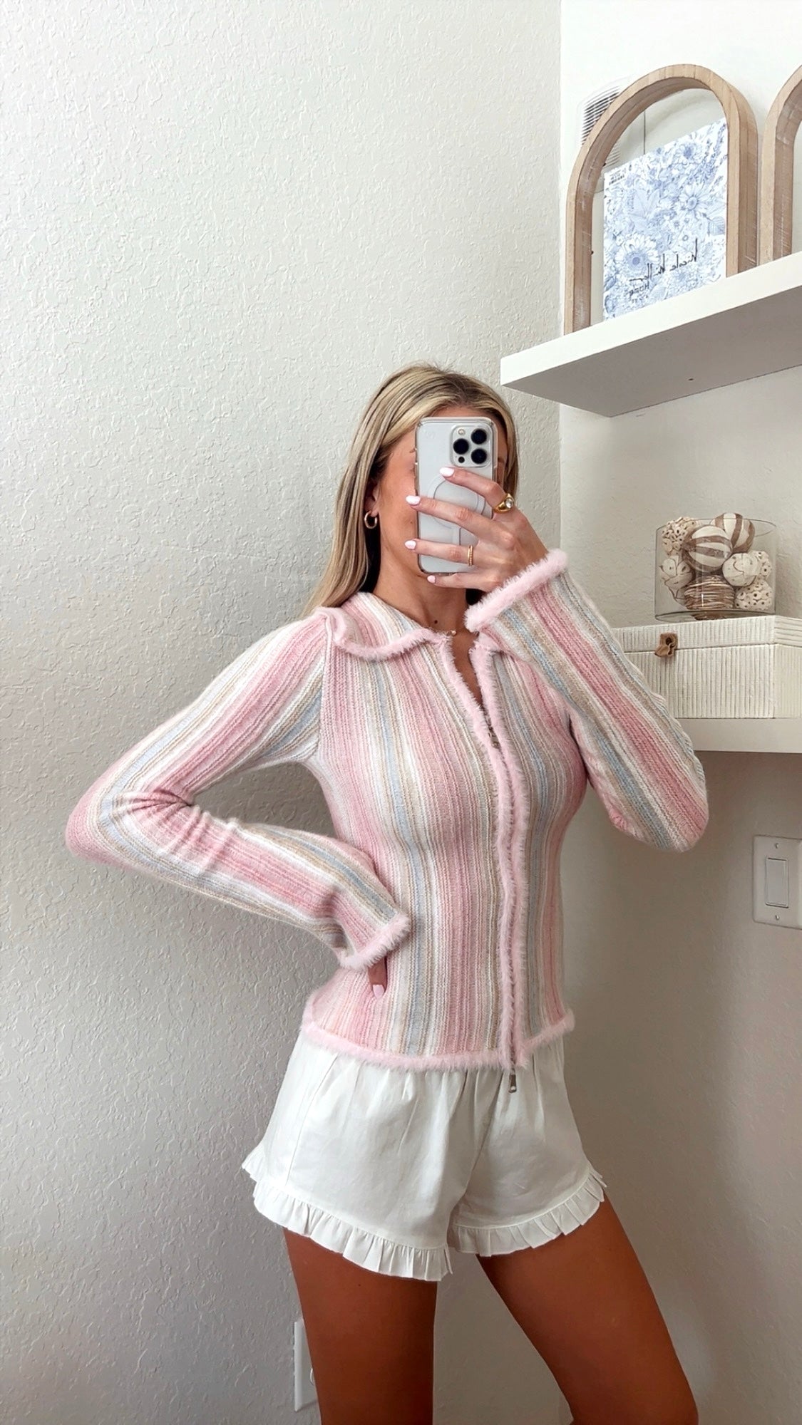 Woman taking a mirror picture wearing a pink pastel striped cardigan and white shorts.