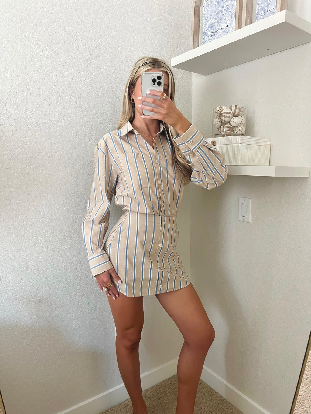 Woman taking a mirror selfie wearing a tan, blue, and white pin striped button down and skort set in a room with shelves and decor.