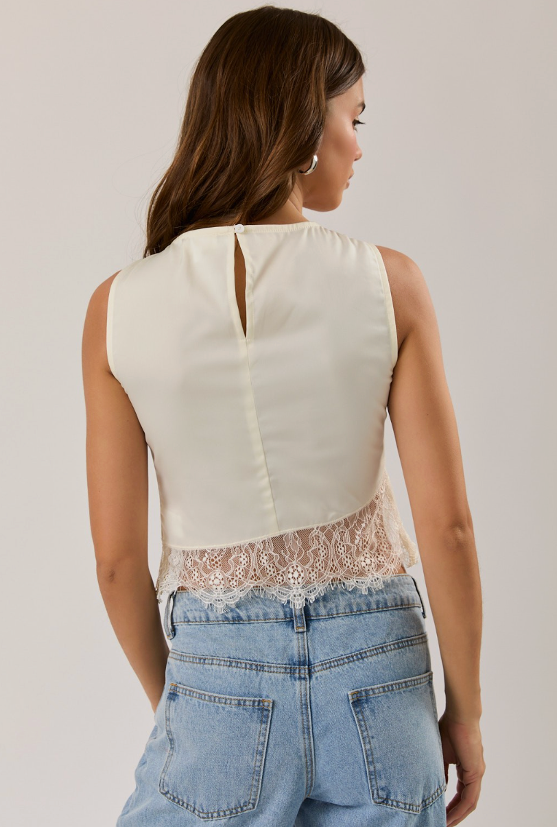 A woman shown from the back wearing a sleeveless cream top with a lace hem and a small keyhole cutout at the back, paired with light blue jeans against a plain light gray studio background.