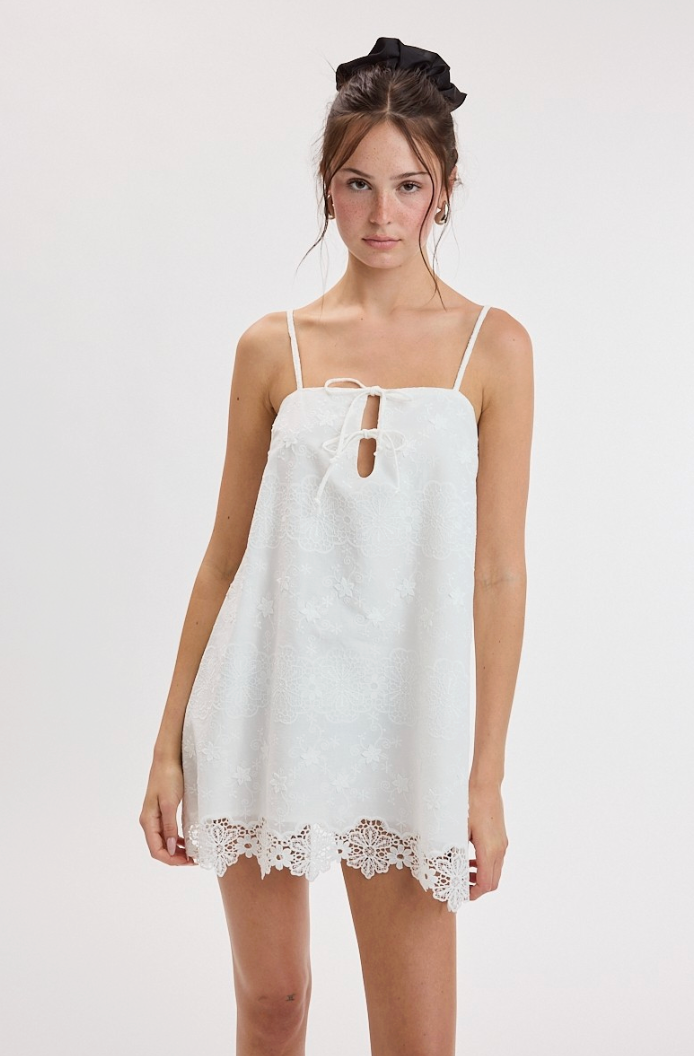 A woman modeling a white sleeveless mini dress with thin straps, a front keyhole cutout, and a lace hem, photographed against a plain light gray studio background.