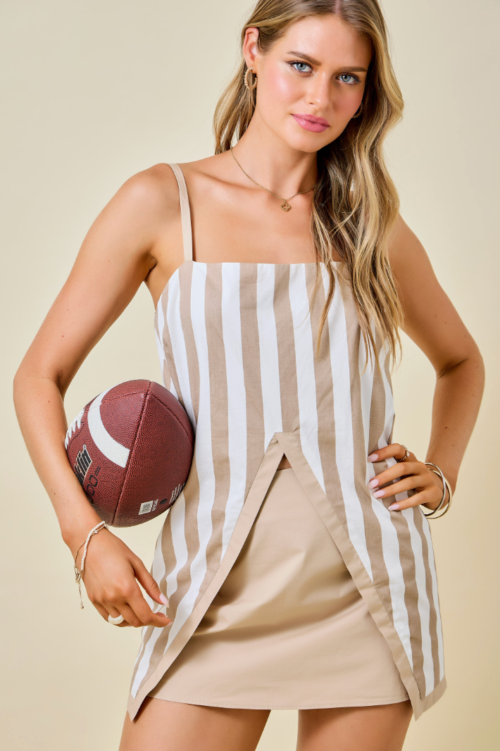 A woman modeling a beige and white vertically striped mini top with a solid beige wrap-style front panel and a tan skort, holding a brown football against a warm beige studio background.