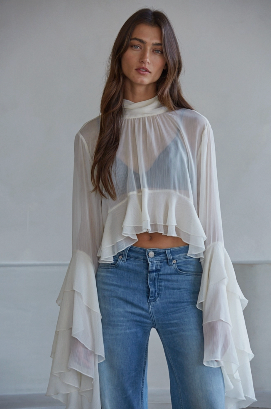Woman wearing an ivory sheer top with jeans against a white wall.