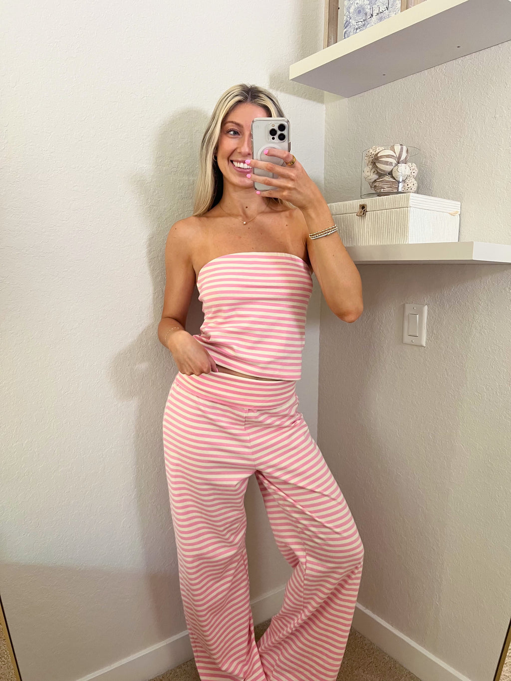 Woman taking a mirror selfie wearing a pink and ivory striped tube top and pants set in a room.