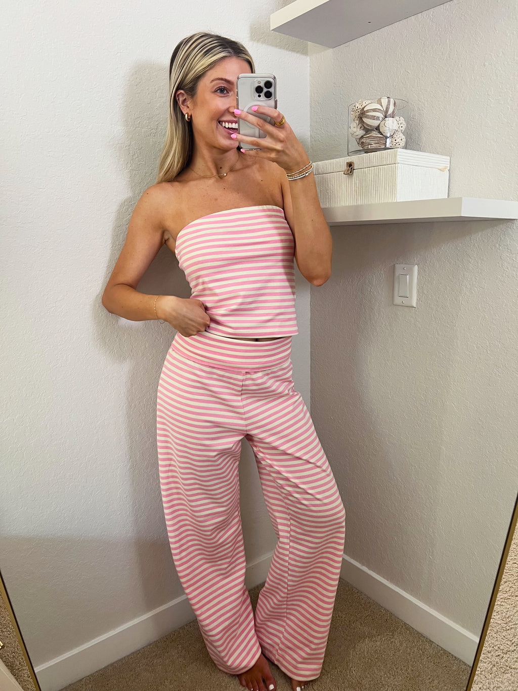 Woman taking a mirror selfie wearing a pink and ivory striped tube top and pants set in a room.