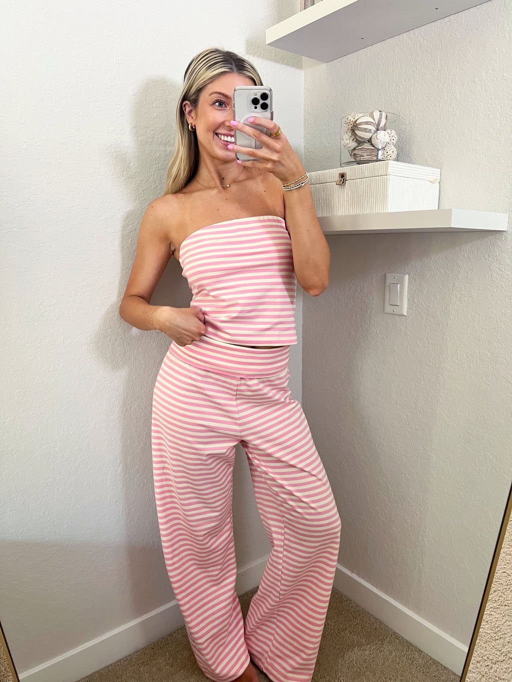 Woman taking a mirror selfie wearing a pink and ivory striped tube top and pants set in a room.