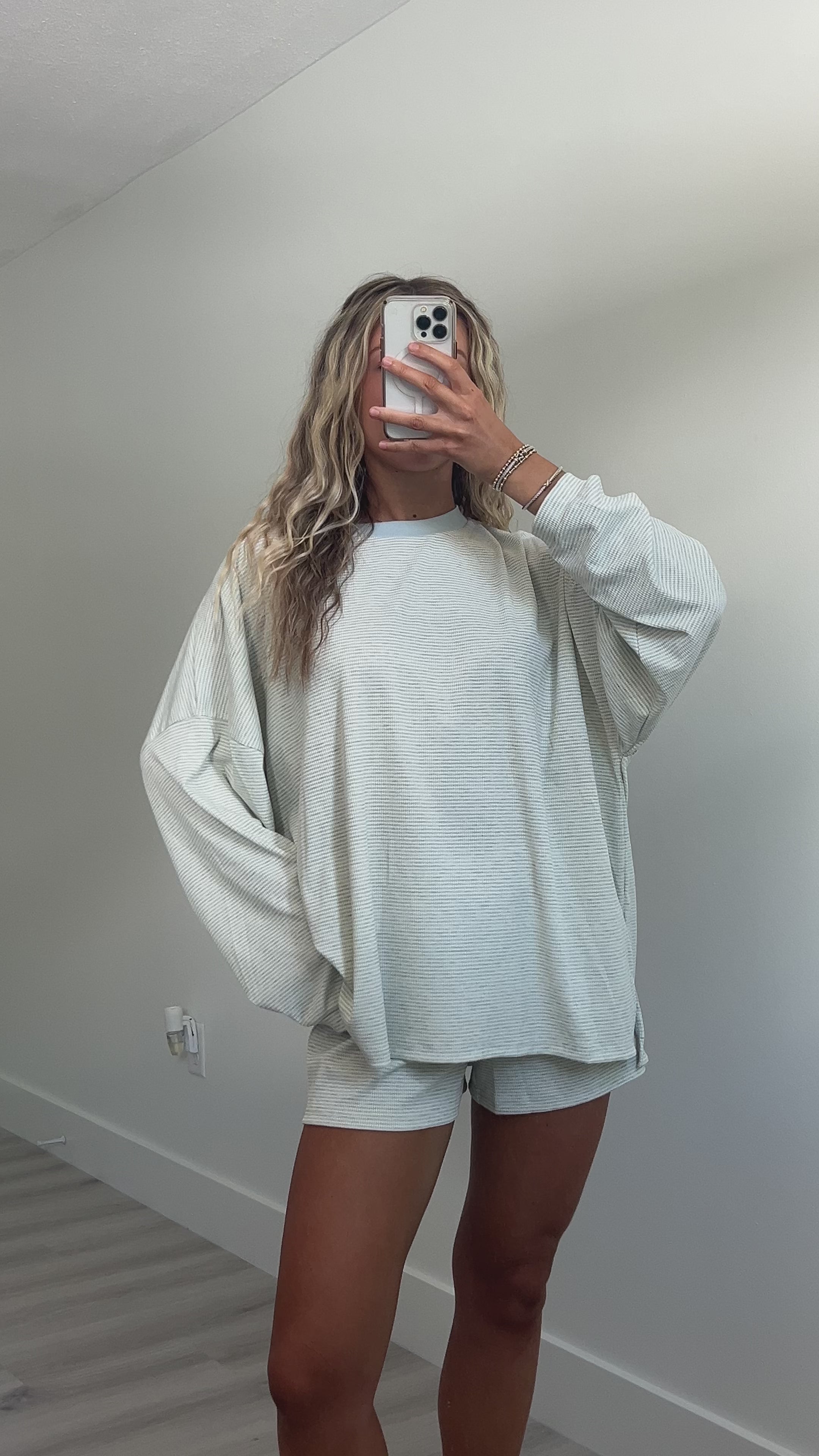 A woman takes a mirror video in a room wearing a light blue striped long-sleeve lounge set consisting of an oversized sweatshirt and matching shorts.